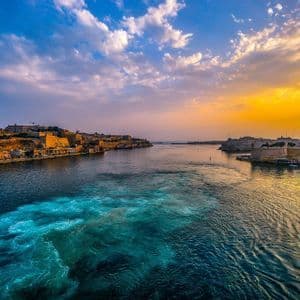 Una vista dall'acqua di un porto storico della città al tramonto, con una grande nave ormeggiata e una scia turbolenta in primo piano.