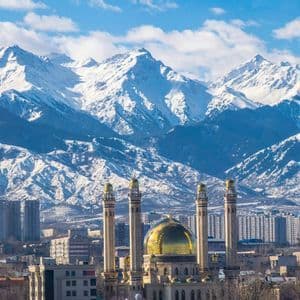 Una moschea con cupola dorata e quattro minareti si erge in un paesaggio urbano ai piedi di una grande catena montuosa innevata.
