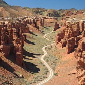 Vue en plongée d'un chemin de terre sinueux traversant un canyon profond avec des formations rocheuses rouges et de la végétation verte.