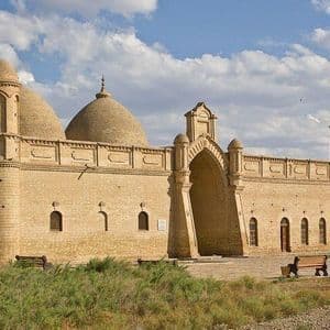 Ein historisches sandfarbenes Gebäude mit mehreren Kuppeln und einem großen Bogengang befindet sich in einer grasbewachsenen Landschaft unter einem bewölkten Himmel.