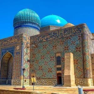 Ein prunkvolles Mausoleum mit zwei blauen Kuppeln und kunstvollen blau-grünen Fliesenmosaiken an den Wänden steht unter einem klaren blauen Himmel.