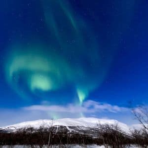 La aurora boreal verde se arremolina en un cielo nocturno estrellado sobre un paisaje montañoso cubierto de nieve.