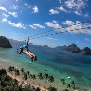 Eine Person gleitet mit einer Seilrutsche über einen tropischen Strand mit Palmen, türkisfarbenem Wasser und entfernten Inseln.