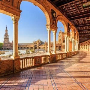 Una vista da un colonnato curvo con archi in mattoni e pilastri in pietra che si affaccia su una piazza soleggiata e un edificio ornato.