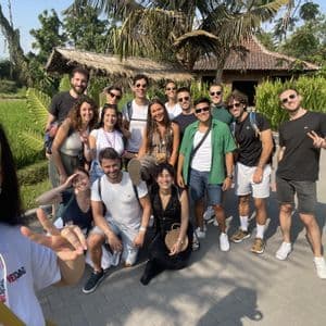 Un gruppo WeRoad in viaggio posa per un selfie sorridente all'aperto, con palme e una capanna tradizionale sullo sfondo.