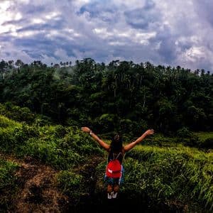 Una donna con uno zaino WeRoad rosso, a braccia aperte su una collina erbosa, osserva una fitta foresta verde sotto un cielo nuvoloso.