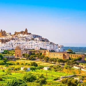 A panoramic view of a whitewashed town on a green hill overlooking the sea under a clear blue sky.