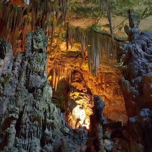 The interior of a large cave with numerous stalactites and stalagmites illuminated by warm, artificial lights.