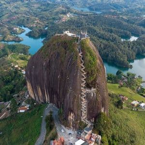 Veduta aerea di un enorme monolito con una scalinata incorporata nel suo fianco, immerso in un paesaggio di verdi colline e laghi blu serpeggianti.