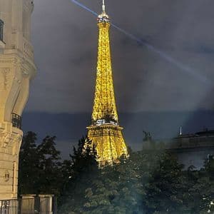 La Torre Eiffel illuminata da luci dorate di notte, il suo faro che proietta un fascio nel cielo nuvoloso, visibile sopra le cime degli alberi.