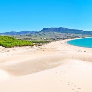 Vue plongeante de dunes de sable blanc menant à une plage incurvée avec de l'eau turquoise, bordée par une forêt verdoyante et des montagnes lointaines.