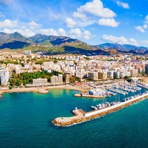 Vista aerea di una città costiera con un grande porto turistico, spiaggia sabbiosa e montagne sullo sfondo sotto un cielo blu e nuvoloso.