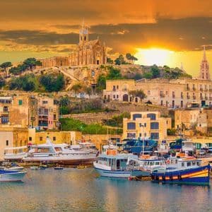 Eine Küstenstadt mit einer Kirche auf einem Hügel überblickt bei Sonnenuntergang einen Hafen voller Boote und Yachten.