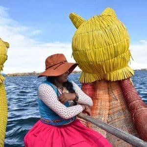 Eine Frau in traditioneller Kleidung und einem braunen Hut sitzt auf einem dekorativen gelben Schilfboot auf einem großen Gewässer.