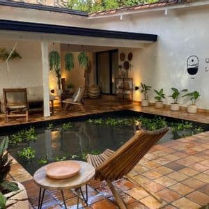 A tiled patio courtyard with a central pond, wooden lounge chairs, a hammock, and many potted plants under warm lighting.
