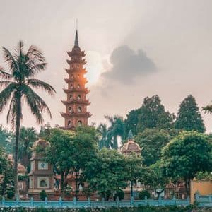 Une pagode à plusieurs niveaux se dresse parmi des arbres verts luxuriants et un palmier alors que le soleil se couche derrière elle.