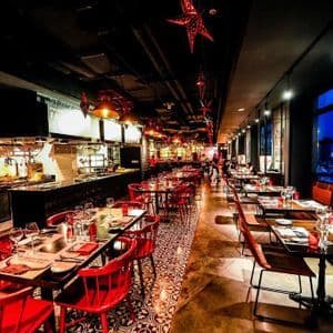 Ein warm beleuchteter Restaurant-Innenraum mit roten Stühlen und gedeckten Tischen an großen Fenstern, die eine nächtliche Stadt-Skyline überblicken.