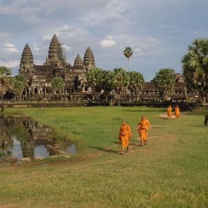 Des moines aux robes orange marchent sur un chemin devant un ancien temple de pierre, qui se reflète dans un étang proche.