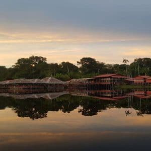 Un villaggio di palafitte su un lago sereno riflette perfettamente la giungla circostante e un cielo al tramonto colorato.