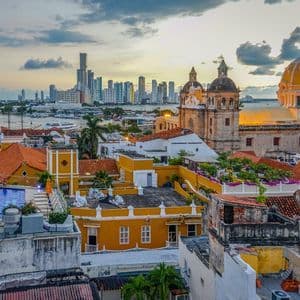 Veduta urbana di edifici storici con tetti in terracotta e una cattedrale, con uno skyline moderno sull'acqua al tramonto.