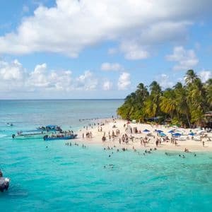Una vista aerea di una spiaggia tropicale affollata con acqua turchese, numerose imbarcazioni e persone che nuotano e prendono il sole sulla sabbia.