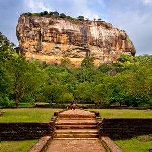 Un sentiero in pietra con gradini conduce attraverso giardini verdi verso un'imponente fortezza rocciosa che si erge sopra una fitta foresta.