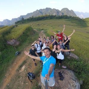 Eine WeRoad-Gruppenreise macht ein lächelndes Selfie auf einem Wanderweg mit Blick auf terrassierte grüne Hügel und ferne Berge.