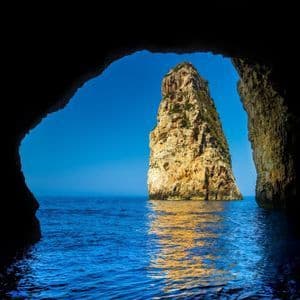 Une haute formation rocheuse ensoleillée émergeant de la mer d'un bleu profond, vue depuis l'embouchure d'une grotte sombre.