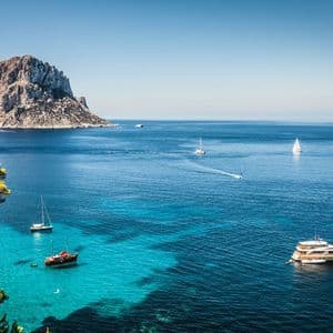 Yachten und Segelboote auf einem ruhigen Meer mit türkisfarbenem und tiefblauem Wasser und einer großen Felseninsel im Hintergrund.