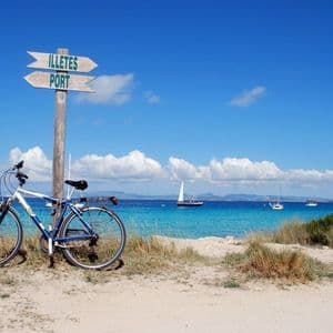 Ein Fahrrad steht neben einem hölzernen Wegweiser an einem Sandstrand mit Blick auf ein türkisfarbenes Meer mit Segelbooten.