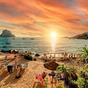 Una vista di una spiaggia affollata durante un vivace tramonto arancione, con persone che nuotano nel mare e barche che navigano vicino a formazioni rocciose distanti.