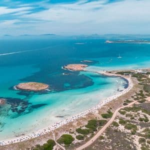 Una vista aerea di una lunga spiaggia di sabbia curva, affollata di gente, che lambisce un mare turchese con barche e piccole isole.