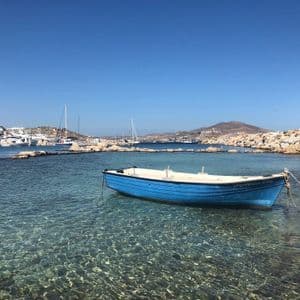 Una piccola barca blu galleggia in acque limpide e turchesi vicino a una costa rocciosa, con un porto e una città in lontananza sotto un cielo azzurro.