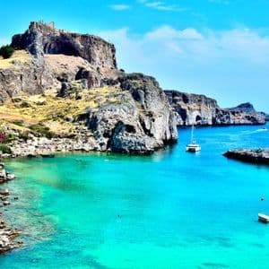 Una scogliera rocciosa sormontata da una fortezza si affaccia su una baia con acqua turchese vibrante dove sono ormeggiate diverse piccole imbarcazioni.