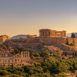 L'antico tempio del Partenone sulla collina dell'Acropoli, illuminato dalla luce dorata del tramonto, con rovine e alberi verdi sottostanti.