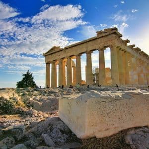An ancient Greek temple with large columns stands on a rocky hill under a blue sky with white clouds at sunset.