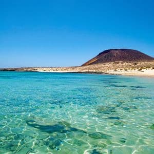 Acqua limpida e turchese lambisce una spiaggia sabbiosa di fronte a una grande collina vulcanica sotto un cielo azzurro senza nuvole.