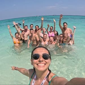 A woman takes a selfie with a WeRoad group trip smiling and waving while standing in clear, turquoise ocean water.
