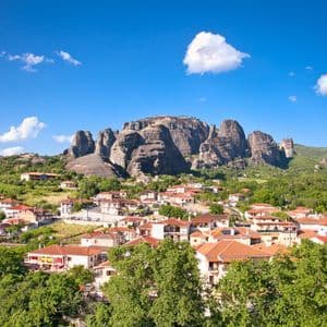 A village with red-tiled roofs is nestled among green trees at the base of towering rock formations under a blue sky.