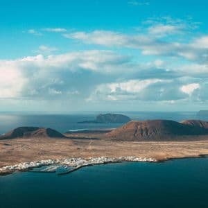 Vue aérienne d'une île volcanique avec un petit village côtier et un port, entourée d'un océan bleu profond sous un ciel partiellement nuageux.
