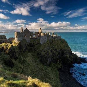Las ruinas de un castillo de piedra se alzan sobre un acantilado verde con vistas al océano bajo un cielo azul con nubes blancas.
