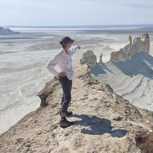 Una persona con gorra se encuentra en una estrecha cresta rocosa, señalando un vasto valle desértico soleado con formaciones rocosas blancas.