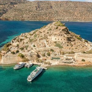Aerial view of a historic fortified island with stone ruins, with several boats docked in the surrounding turquoise water.