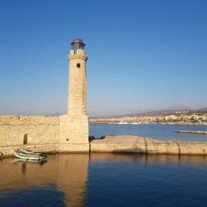 A tall stone lighthouse stands on a seawall in a harbor, with a city and mountains visible in the background under a clear blue sky.