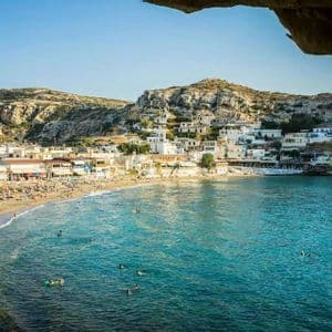 Una vista desde un saliente rocoso de una playa concurrida con gente nadando en agua turquesa frente a un pueblo costero.