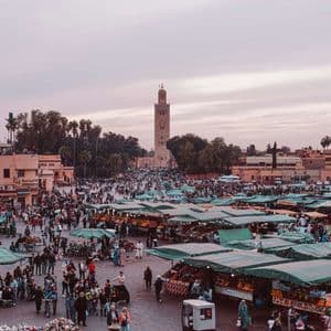 Una bulliciosa plaza de mercado llena de gente y puestos con toldos verdes, dominada por un alto minarete bajo un cielo crepuscular rosado.