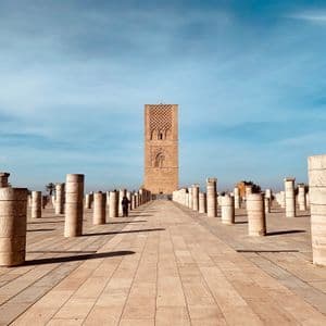 Des rangées de colonnes de pierre emplissent une vaste place pavée, menant à une haute tour historique sous un ciel bleu.