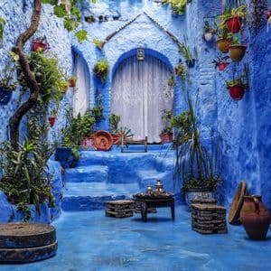 Ein Innenhof mit strukturierten blauen Wänden, Stufen und Boden, dekoriert mit Topfpflanzen, Steinhockern und einem kleinen Tisch mit Teeservice.