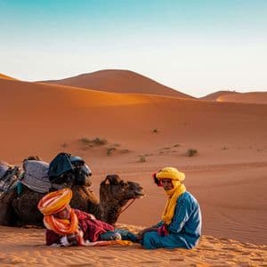Zwei Männer in farbenfrohen Turbanen ruhen sich mit ihren gesattelten Kamelen auf einer Sanddüne in einer weiten Wüstenlandschaft aus.