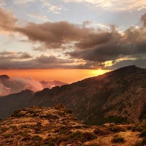 Il sole tramonta dietro una catena montuosa scoscesa, proiettando un bagliore arancione su un cielo nuvoloso e sulle valli sottostanti.